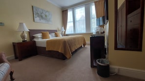 A hotel room with a bed, bedside lamps, a painting, a window with curtains, a desk, and a trash bin. - Two Beaches (Paignton)