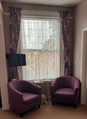 A room with two purple armchairs, a small table with a laptop, a television, and a window with floral curtains.