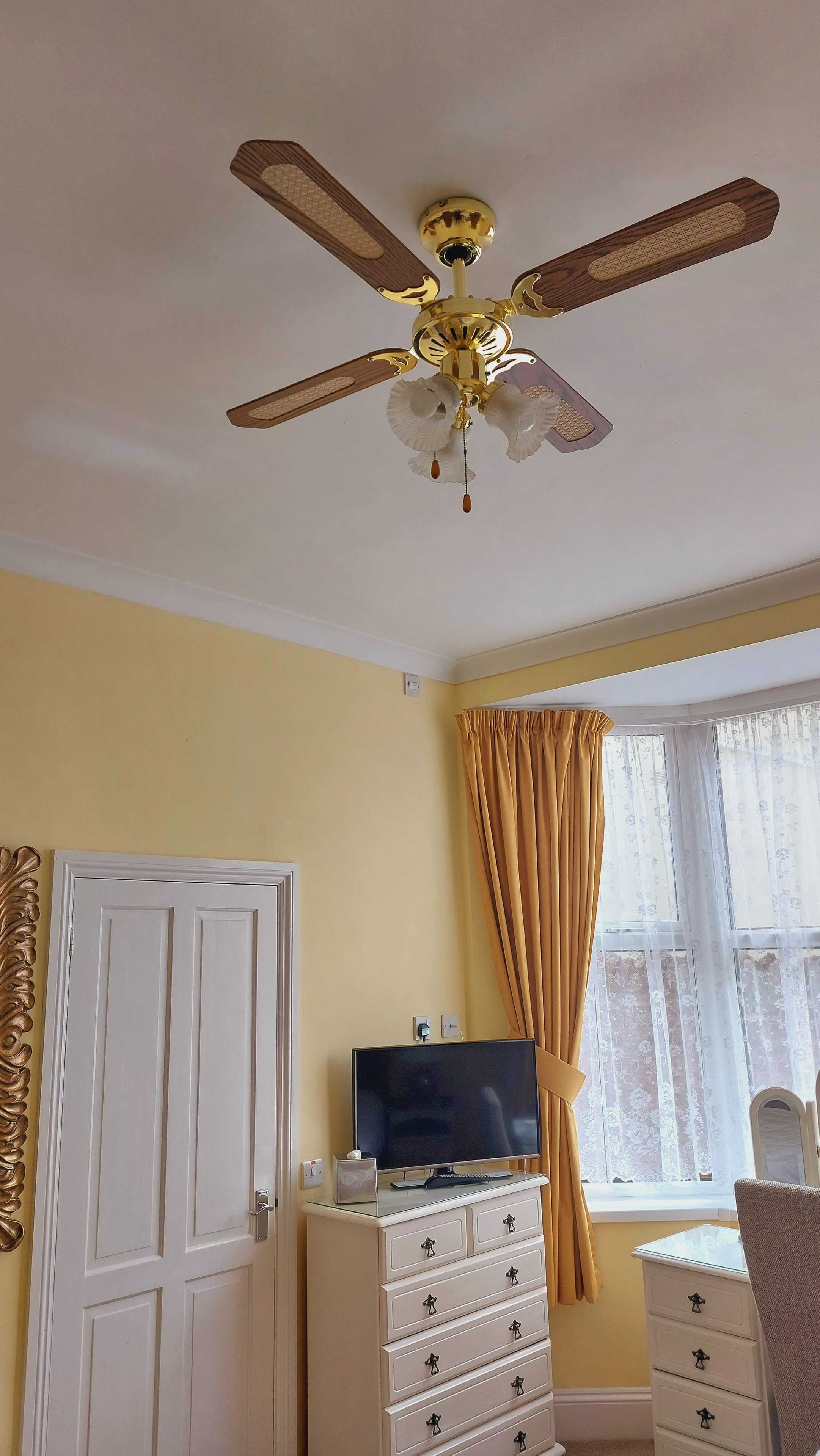 Una habitación con ventilador de techo, televisor sobre una cómoda, una puerta y una ventana con cortinas.