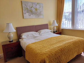 A hotel room with a bed, two bedside lamps, a nightstand, a window with curtains, and a floral painting on the wall.