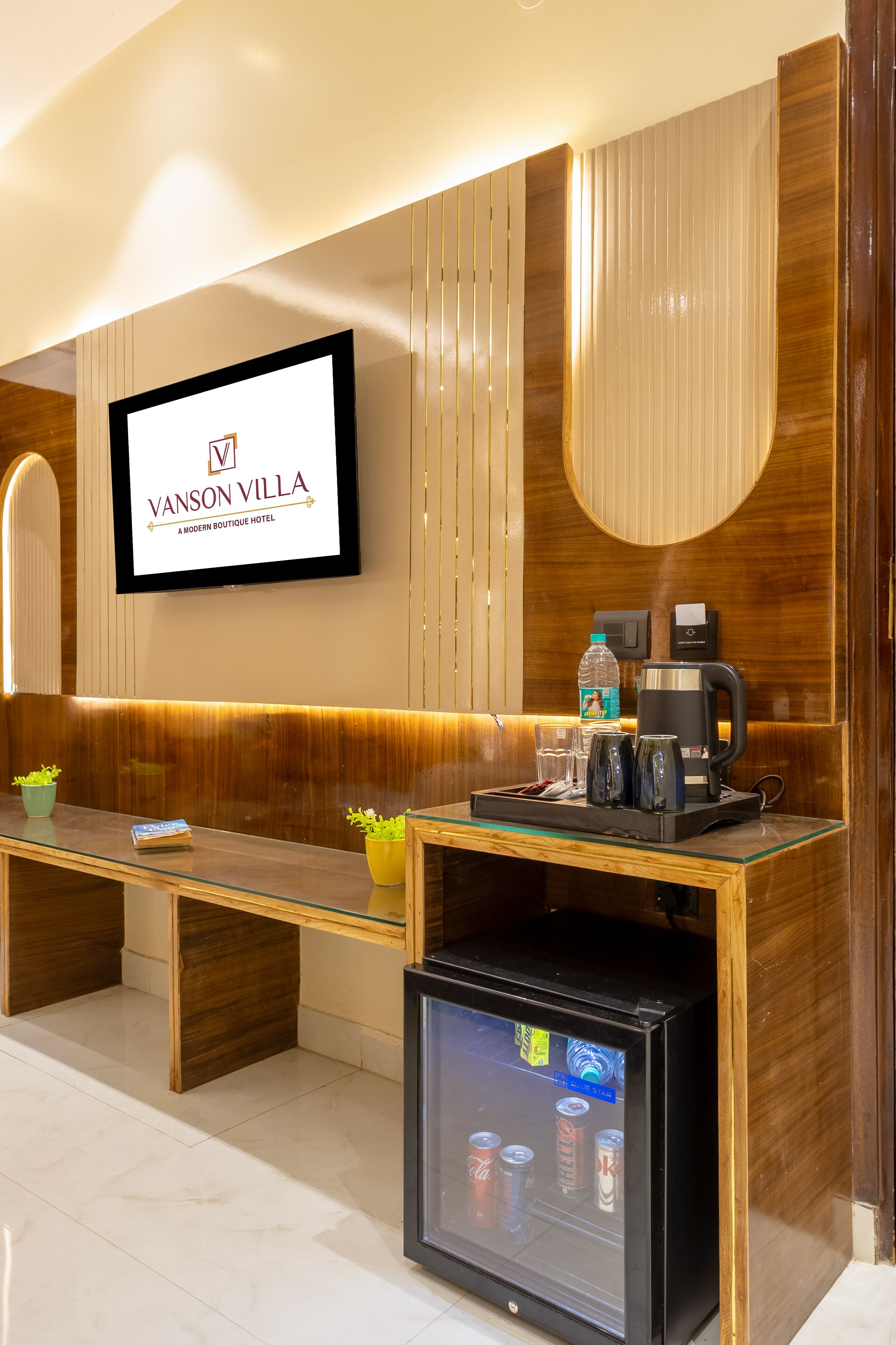 A modern hotel lobby with a reception desk, a coffee machine, a refrigerator stocked with beverages, and a television displaying the hotel name.