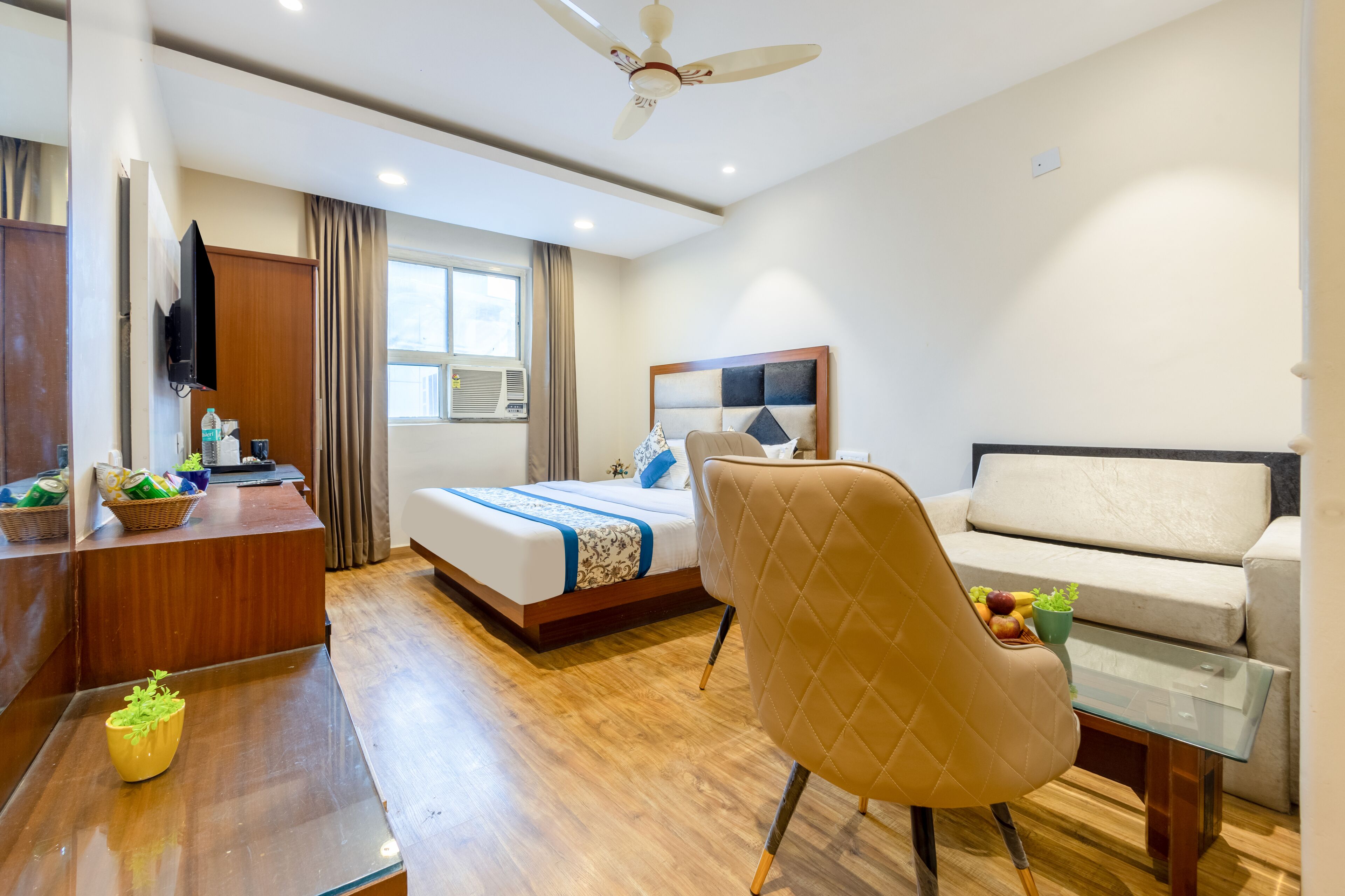 A hotel room with a bed, a sofa, a desk, a chair, a ceiling fan, and a window.