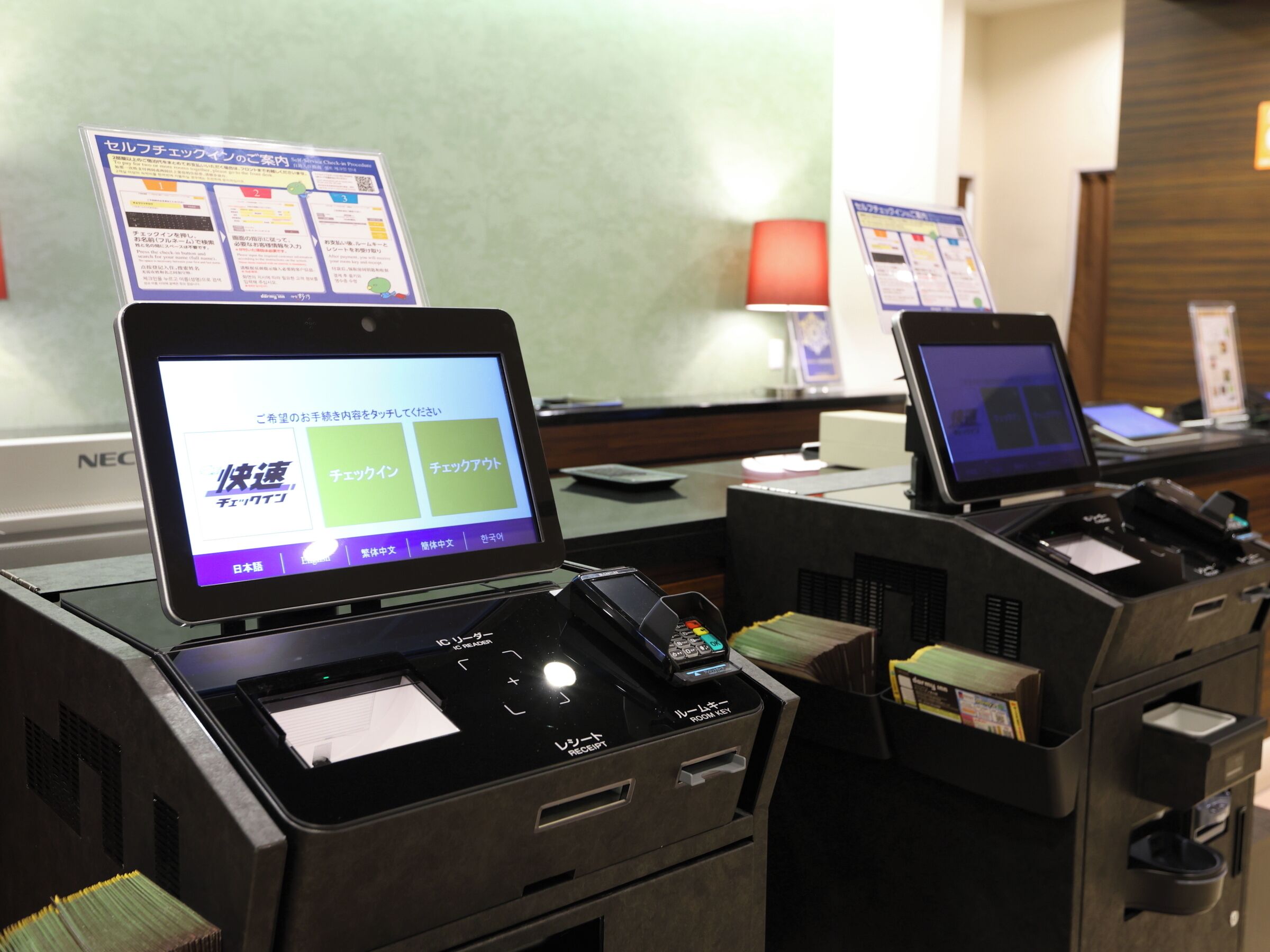 Un mostrador de recepción con dos quioscos de autoservicio que muestran un menú en japonés.