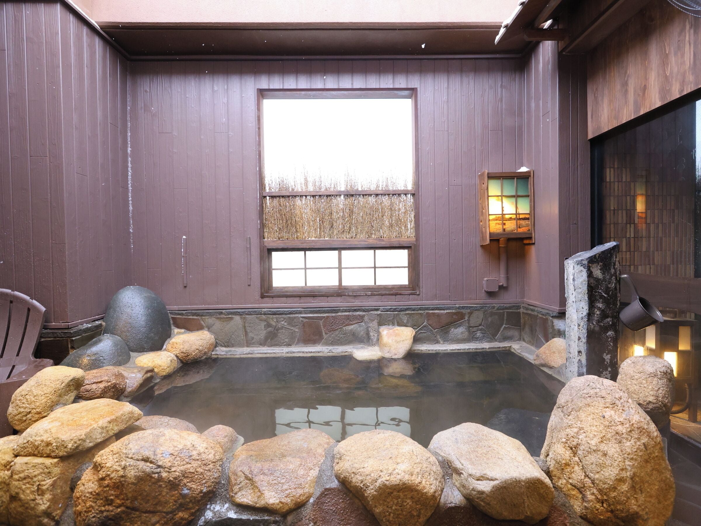 Uma onsen japonesa tradicional com uma nascente quente de pedra, paredes de madeira e vista para o céu através de uma janela.