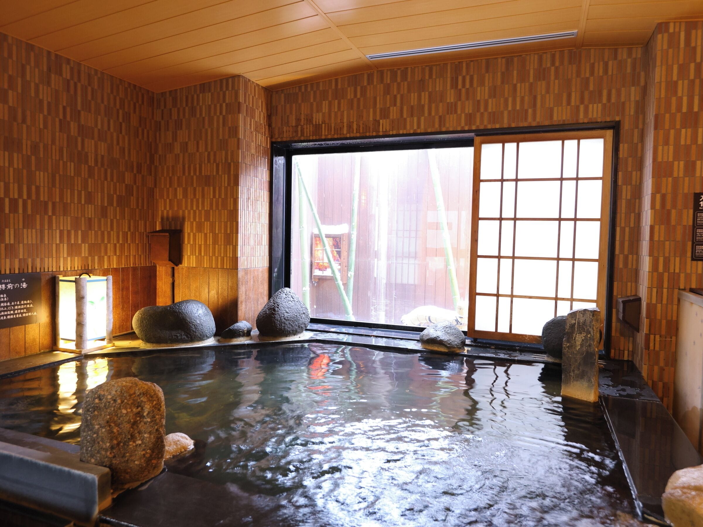 A traditional Japanese onsen with a wooden interior, a large window, and several hot stones in the water.
