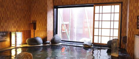 A traditional Japanese onsen with a wooden interior, a large window, and several hot stones in the water.