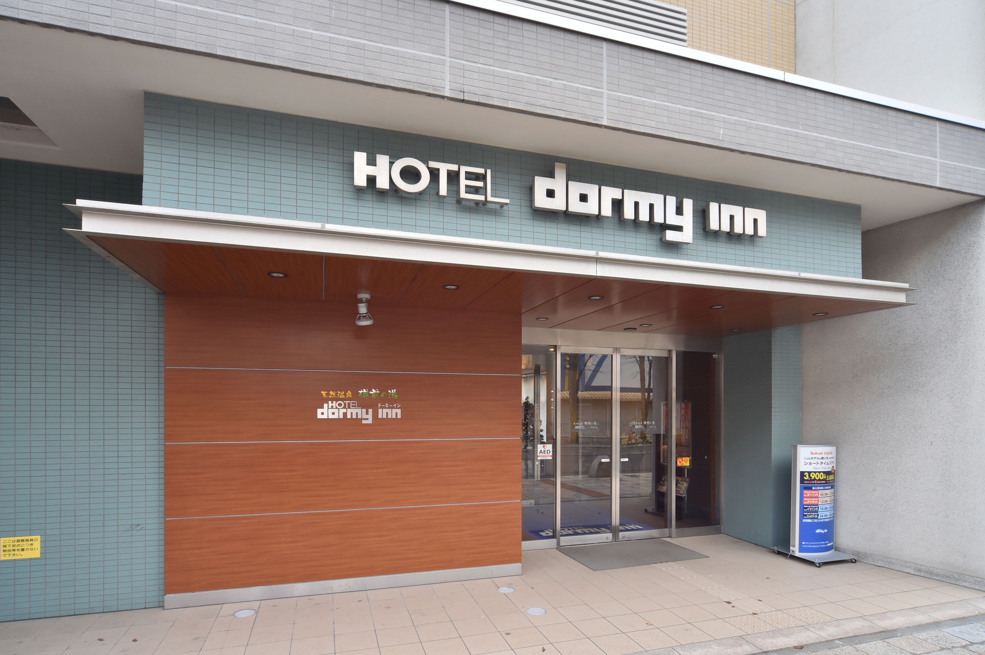 The exterior of Hotel Dormy Inn with a modern facade, large glass entrance, and a prominent sign above the doors.