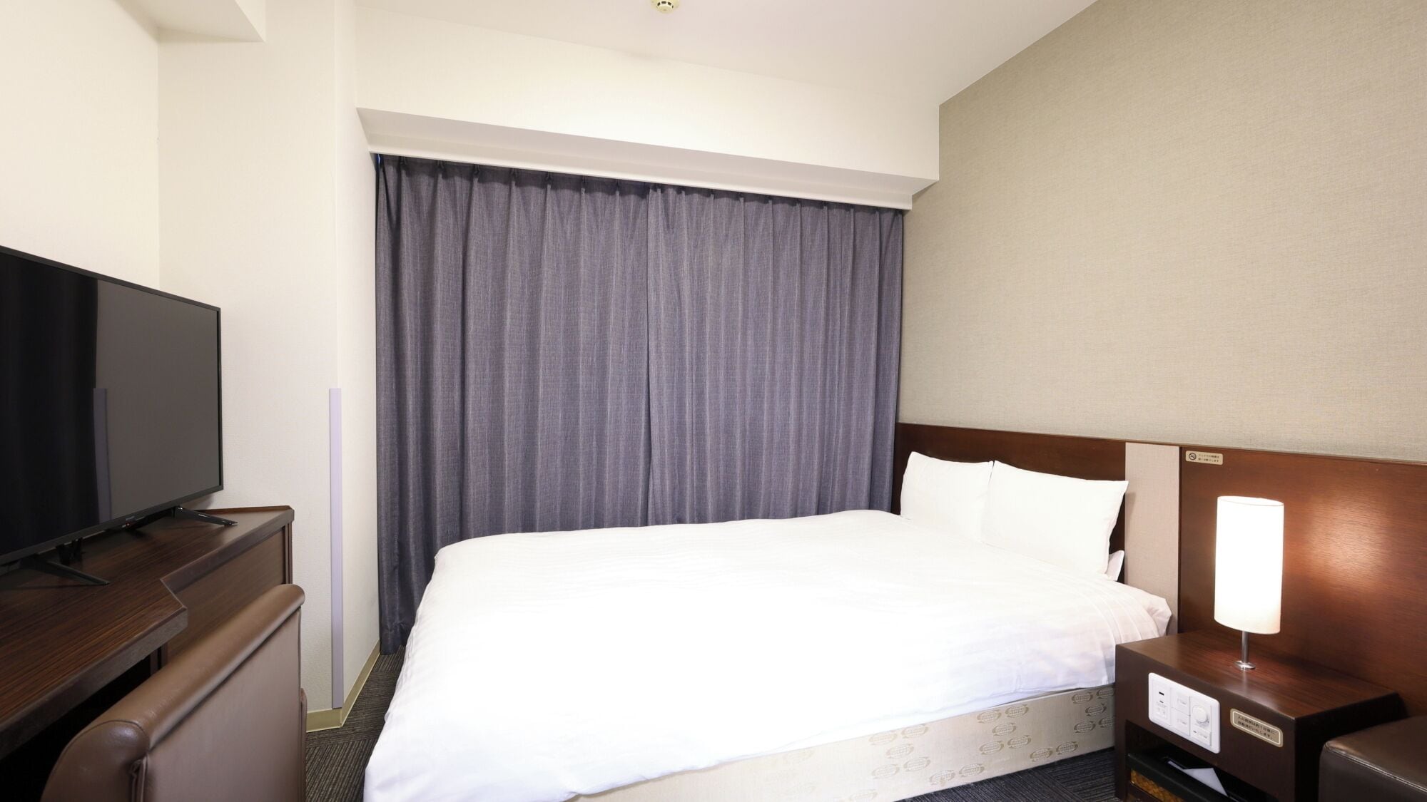Habitación de hotel con cama, televisor sobre una cómoda, mesita de noche con lámpara y un ventanal con cortinas.
