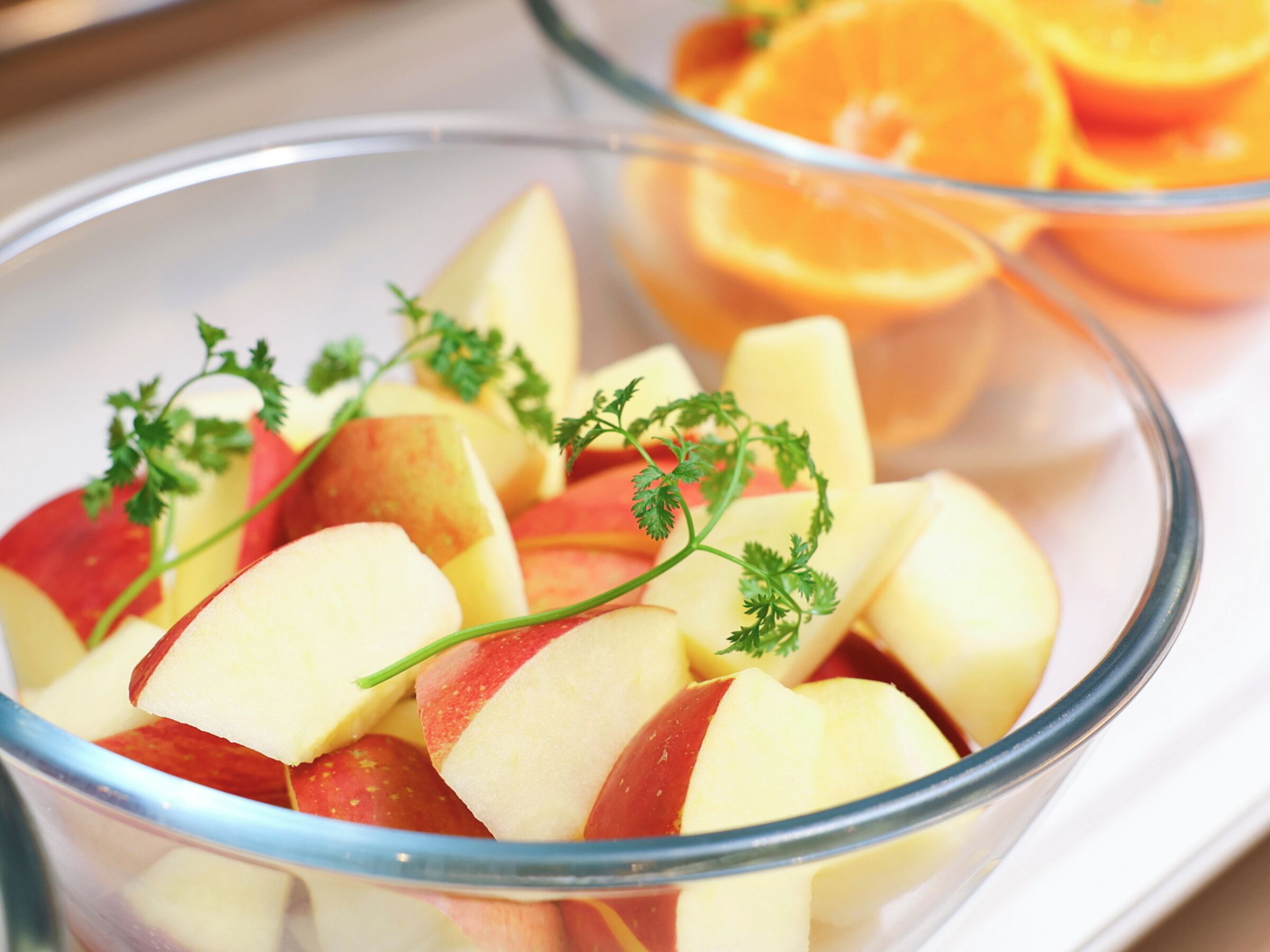 A bowl of sliced apples garnished with parsley, with a glass of orange juice in the background.