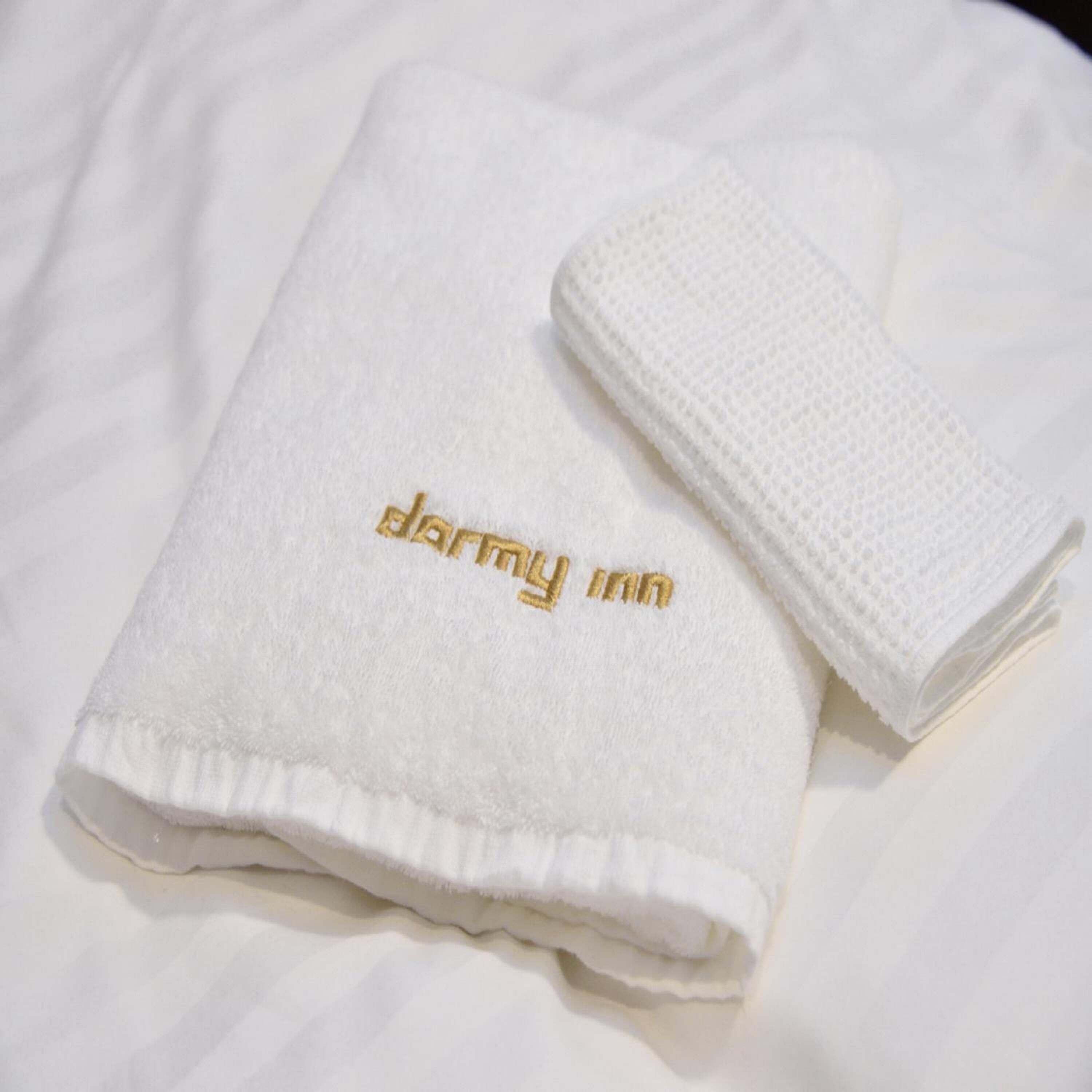 A folded white towel with "darmy inn" embroidered in gold on it, placed on a white bed.