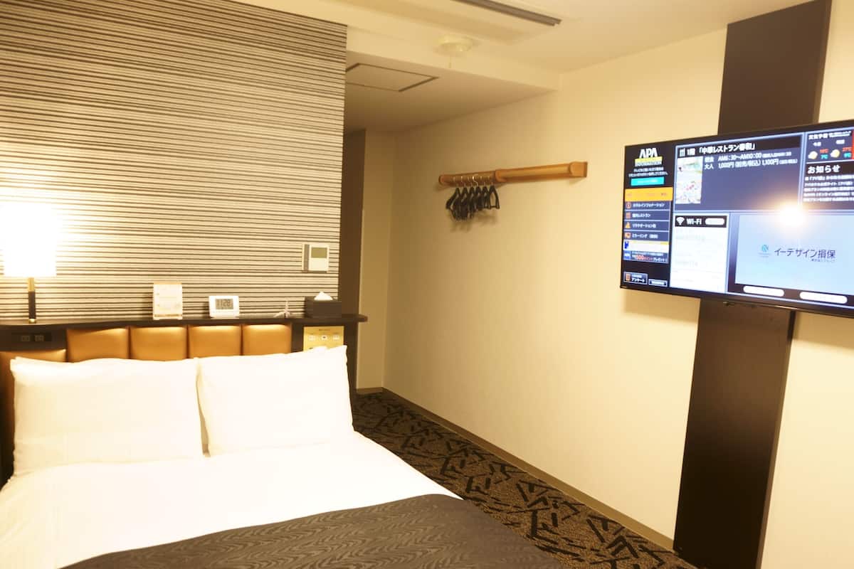 A hotel room with a bed, a television mounted on the wall, and a small table with a clock and a tissue box.