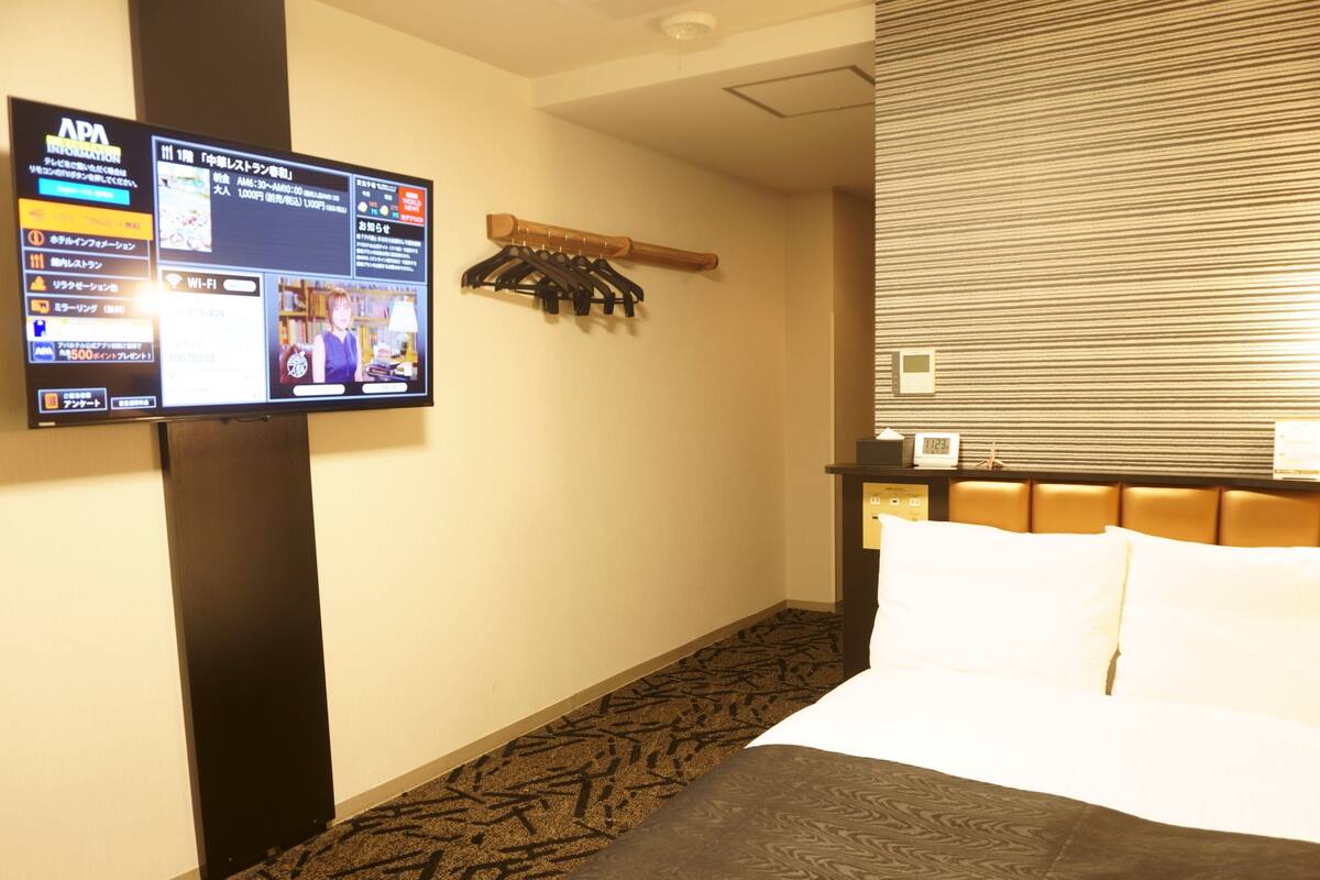 A hotel room with a bed, a TV mounted on the wall displaying a program, a coat rack, and a bedside table with a clock.
