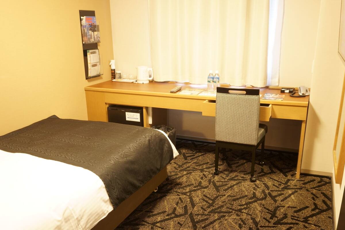A hotel room with a bed, a desk with a chair, a small table with a pitcher and glasses, and a window with curtains.