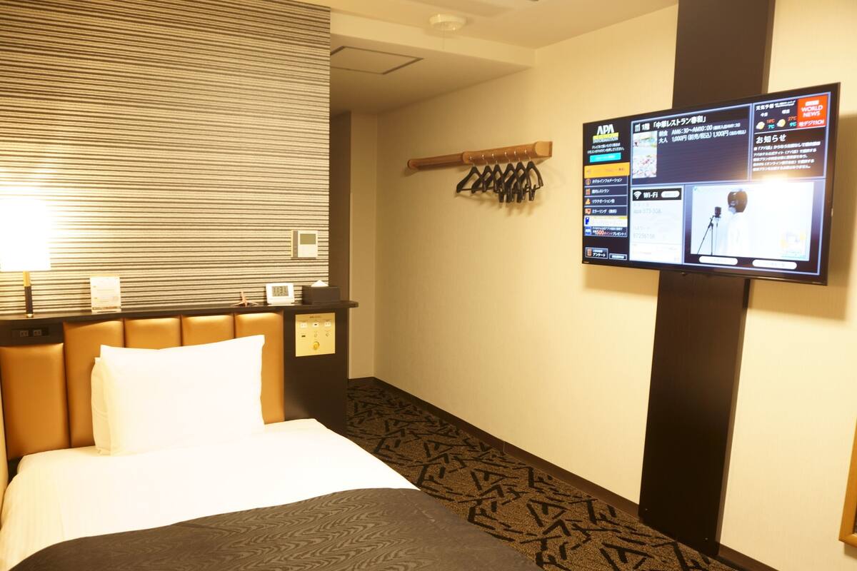 A hotel room with a bed, a control panel, a TV mounted on the wall, and a patterned carpet.