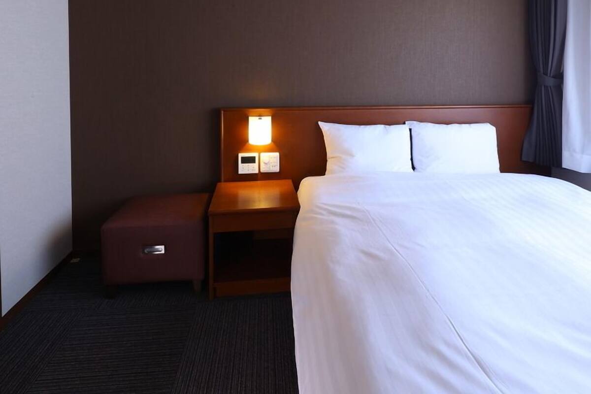 A hotel room with a bed, a nightstand with a lamp, a control panel, and a small drawer.
