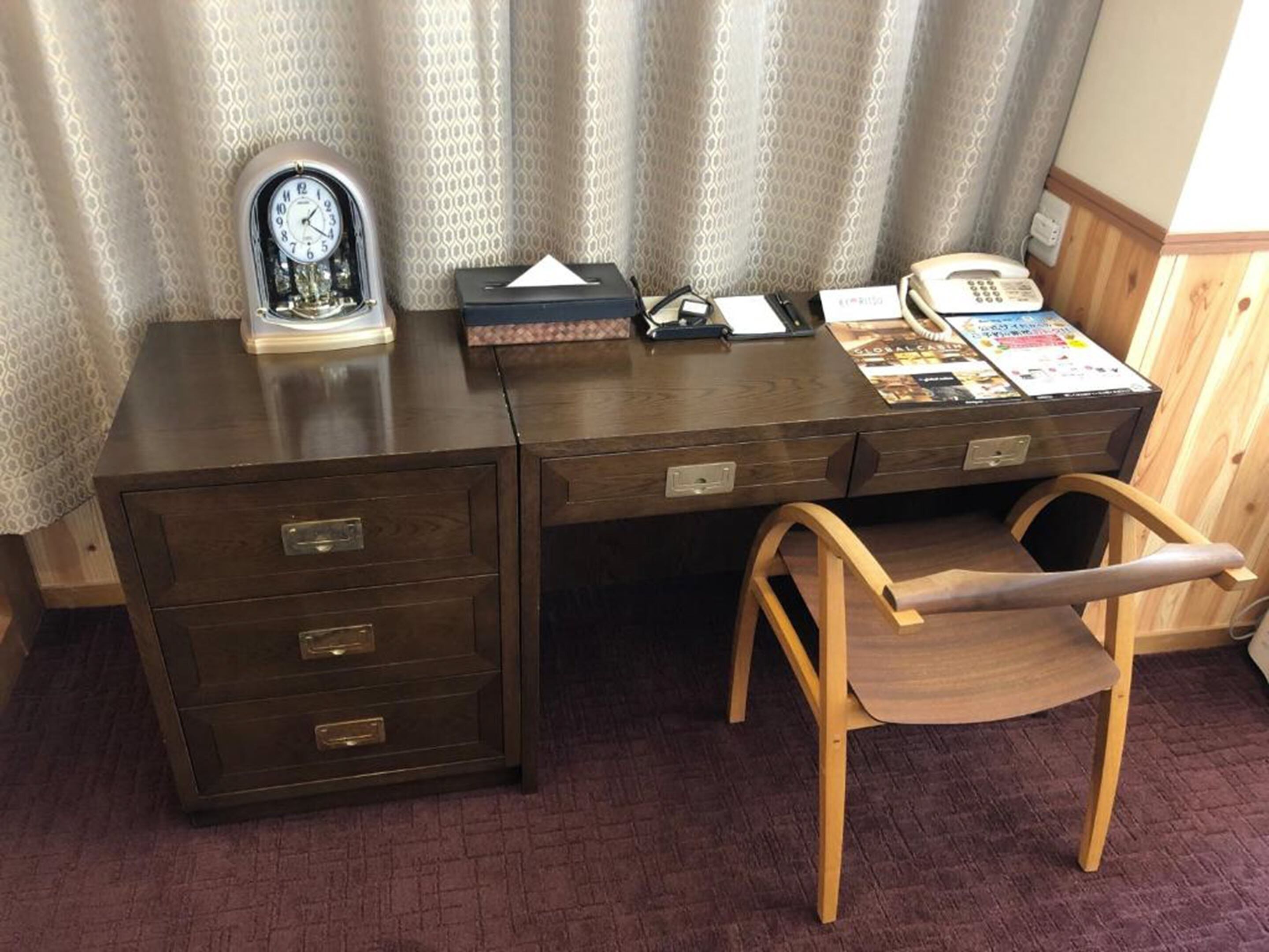 A wooden desk with drawers, a clock, a telephone, and a chair.