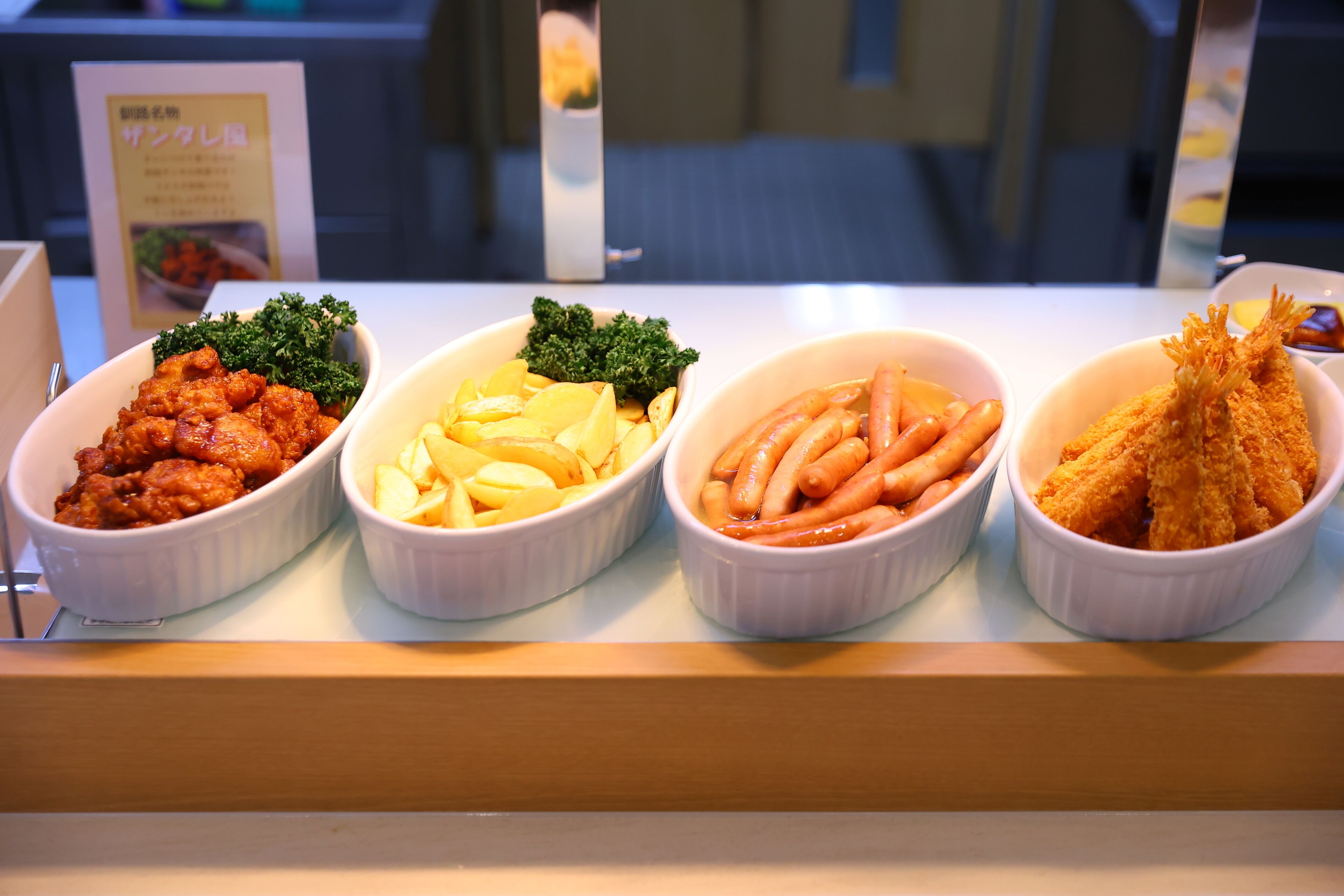 A food display with four dishes: fried chicken, potatoes, sausages, and fried fish.