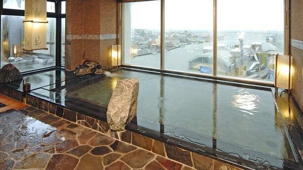 A hot spring area with a stone floor, a large rock, and a view of a port through large windows. - Natural Hot Spring Dormy Inn Premium Kushiro (Kushiro)