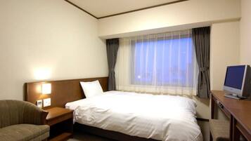 A hotel room with a bed, a desk with a computer, a chair, and a nightstand with a lamp.