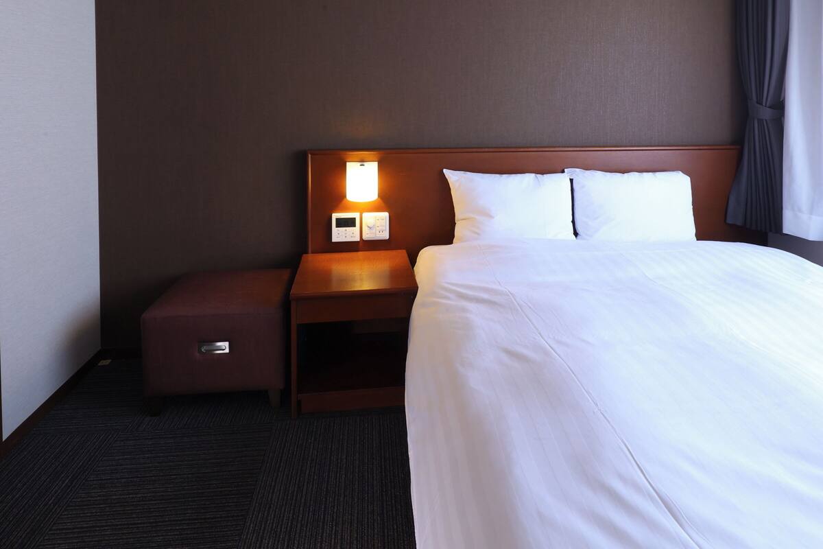 A hotel room with a neatly made bed, a nightstand with a lamp, and a control panel on the wall.