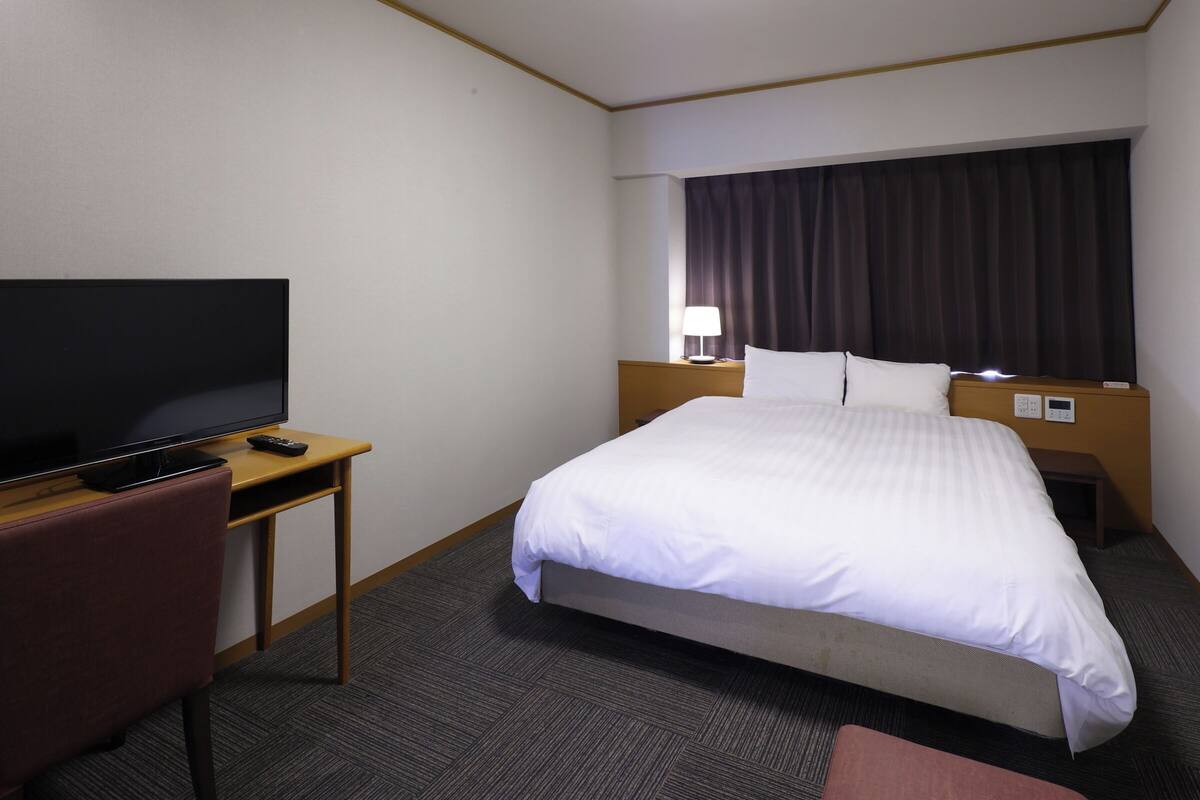 A hotel room with a bed, a desk with a television, a chair, and a nightstand with a lamp.