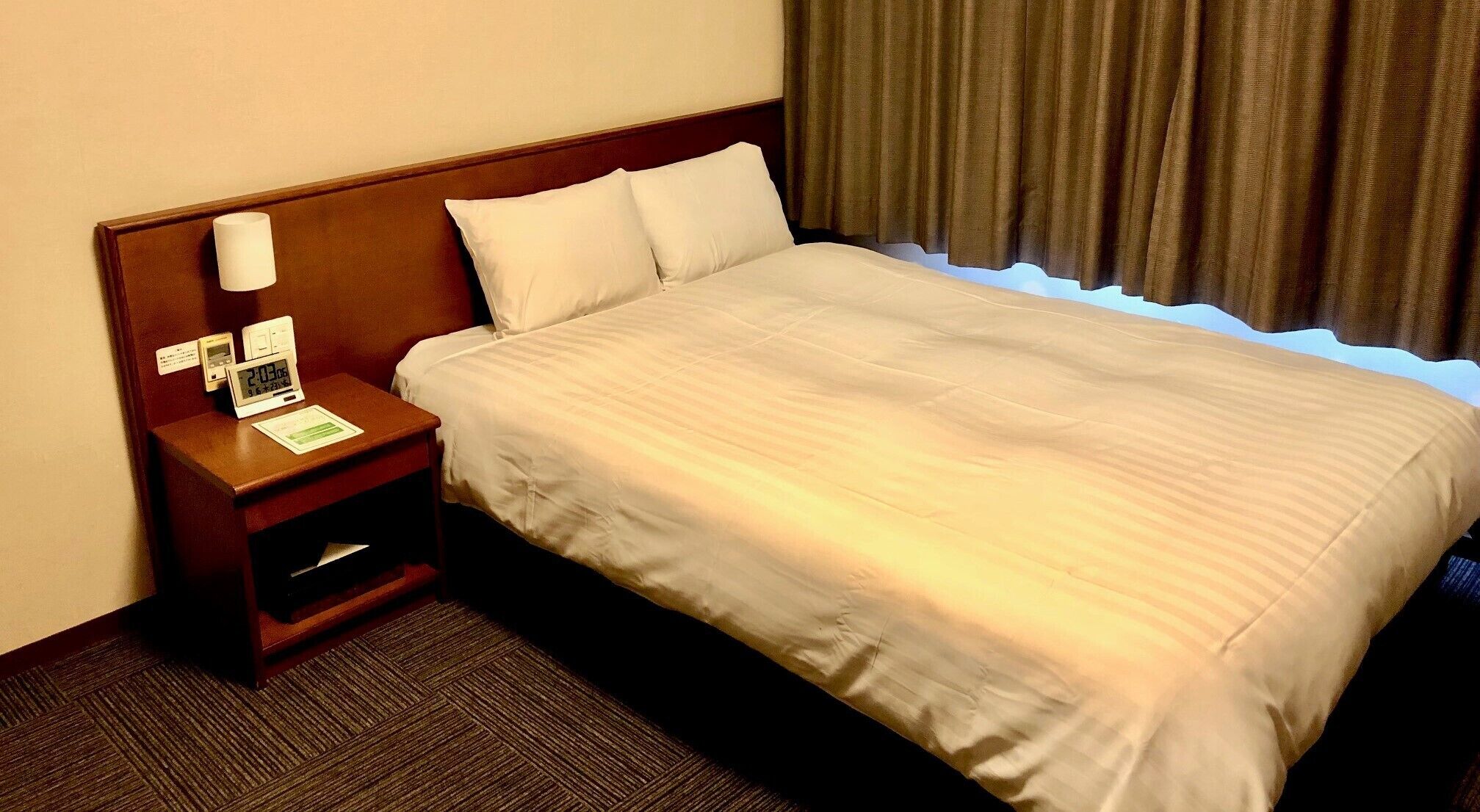 Une chambre d’hôtel avec un lit, une table de chevet avec une lampe, une horloge numérique et un petit tiroir.