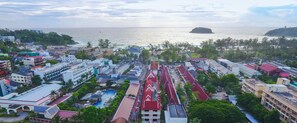 An aerial view of a coastal city with a prominent swimming pool area, buildings with red roofs, and a view of the sea and an island.