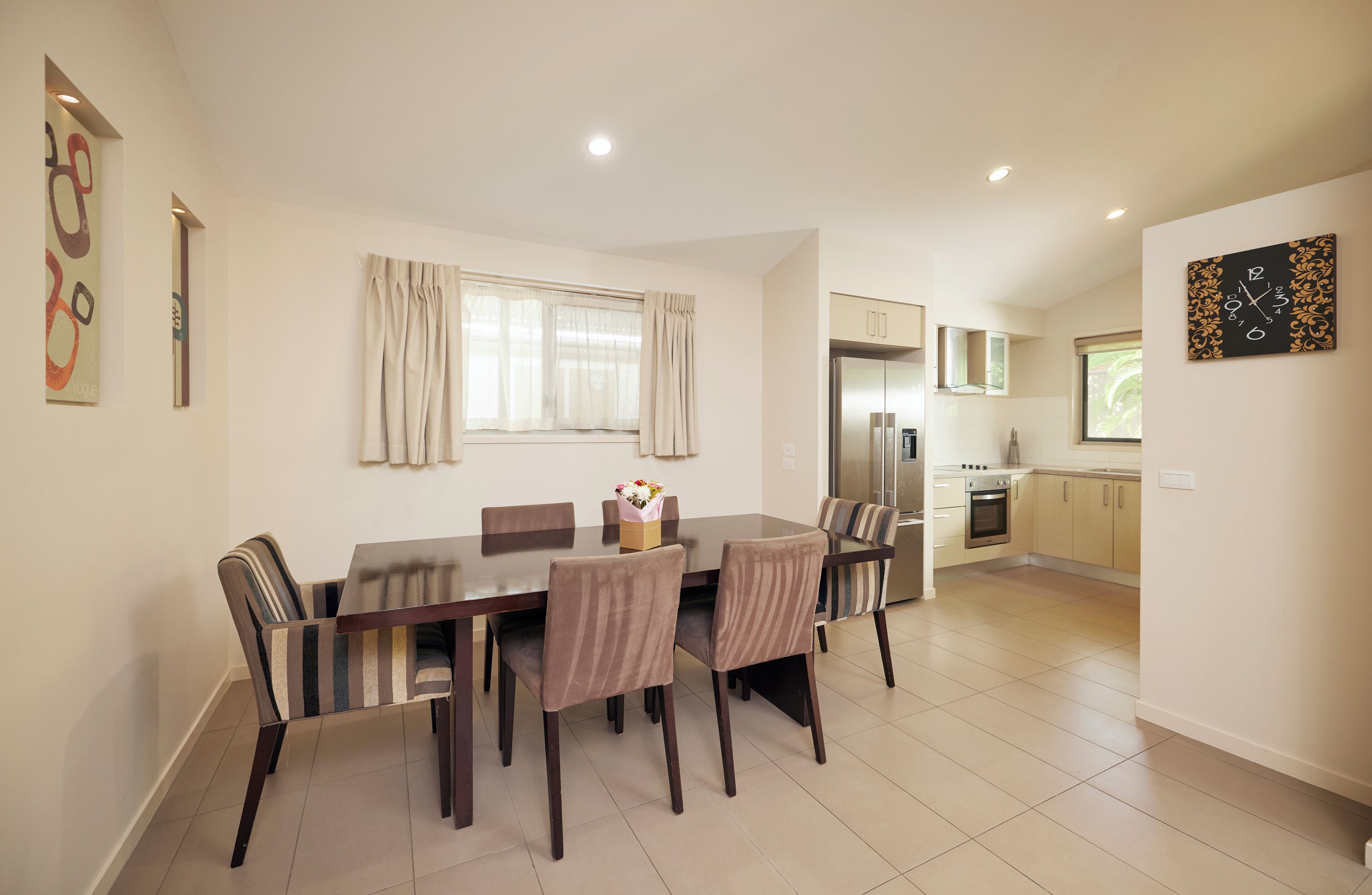 A modern kitchen with a built-in oven and refrigerator, a dining area with a table and chairs, and a window with curtains.