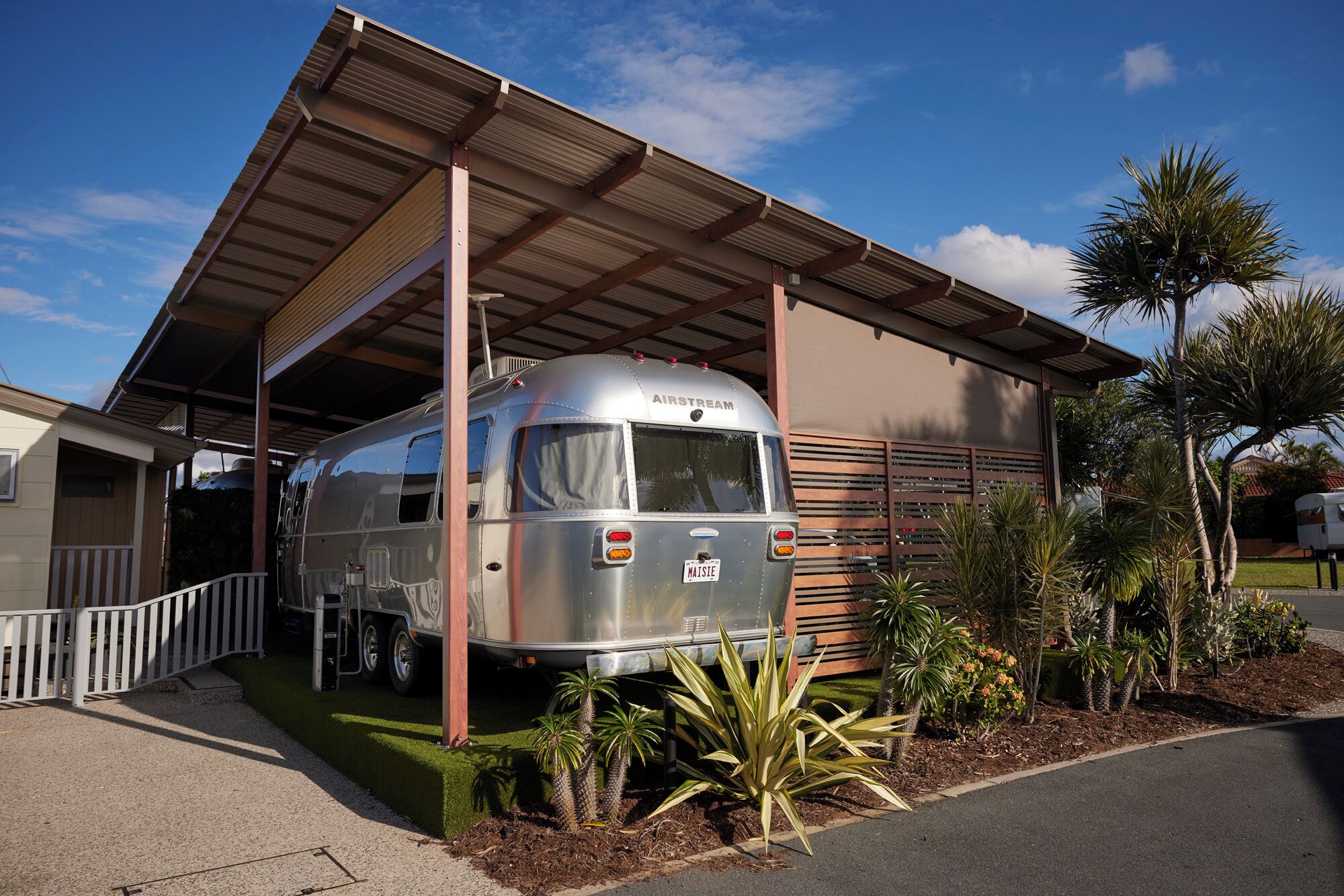 Una caravana Airstream antigua estacionada bajo un techo, con una valla blanca y palmeras.