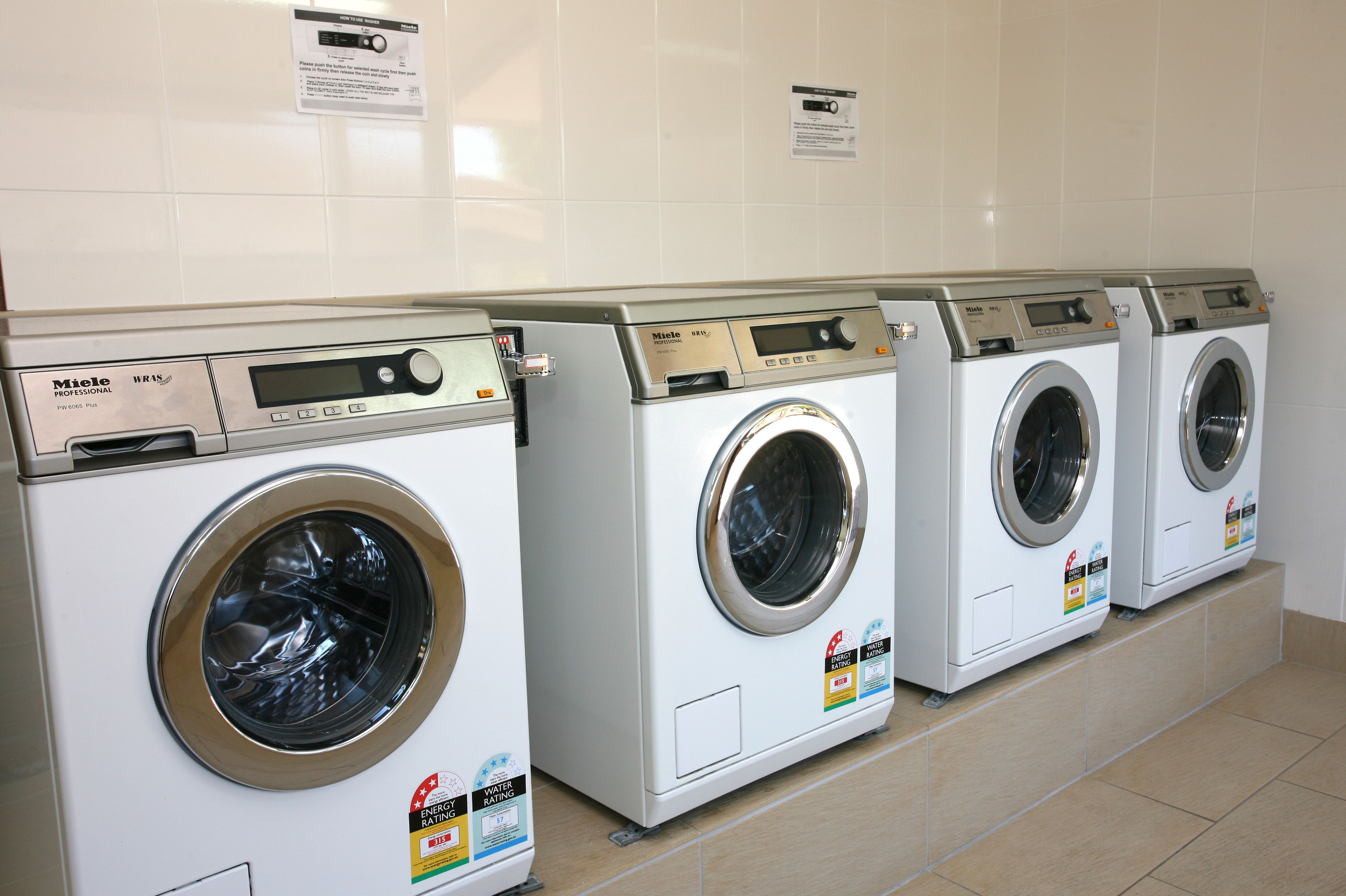 A row of white, professional-grade, front-loading washing machines with digital displays and energy efficiency labels.