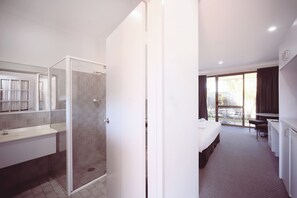 Habitación de hotel moderna con ducha envuelta en vidrio, cama blanca, escritorio y vista al exterior.
