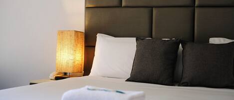 A neatly made bed with white and dark pillows, a white towel, and a bedside lamp.