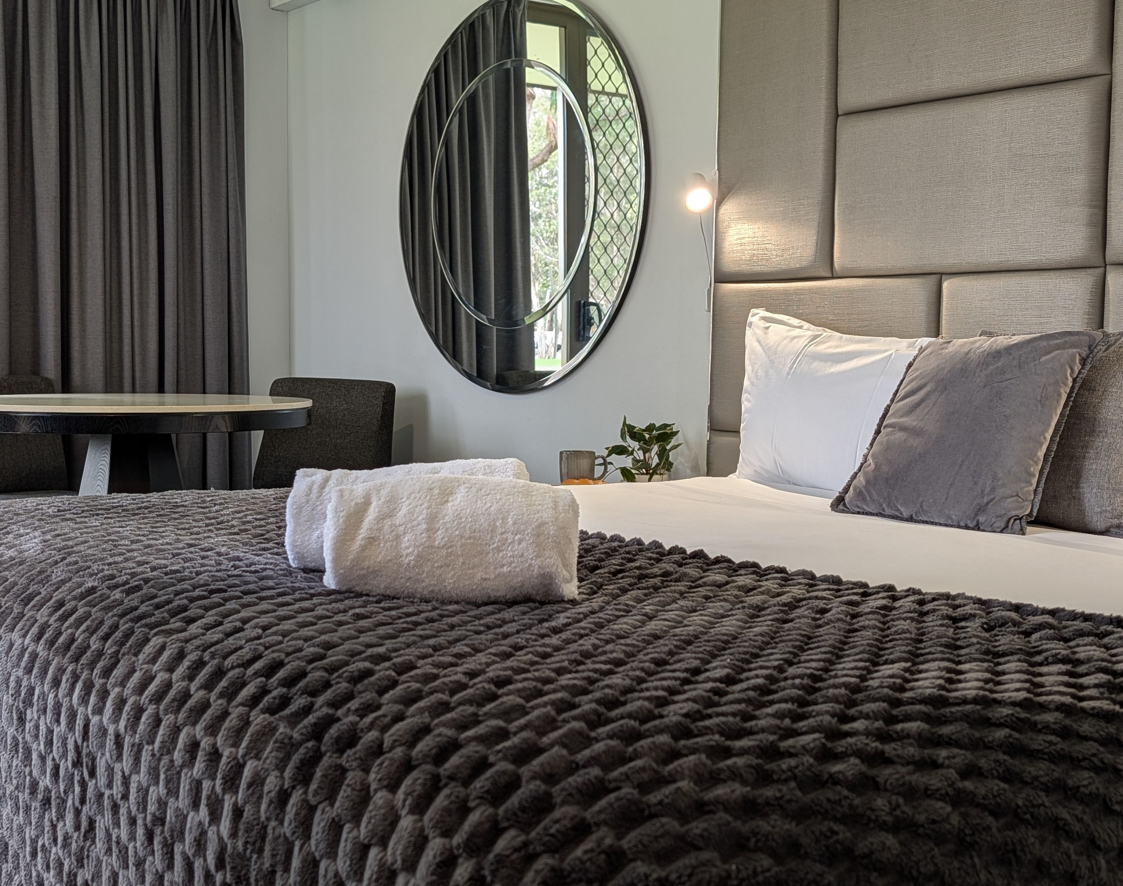 A modern hotel room with a large bed, a round mirror, a small table, and a seating area.