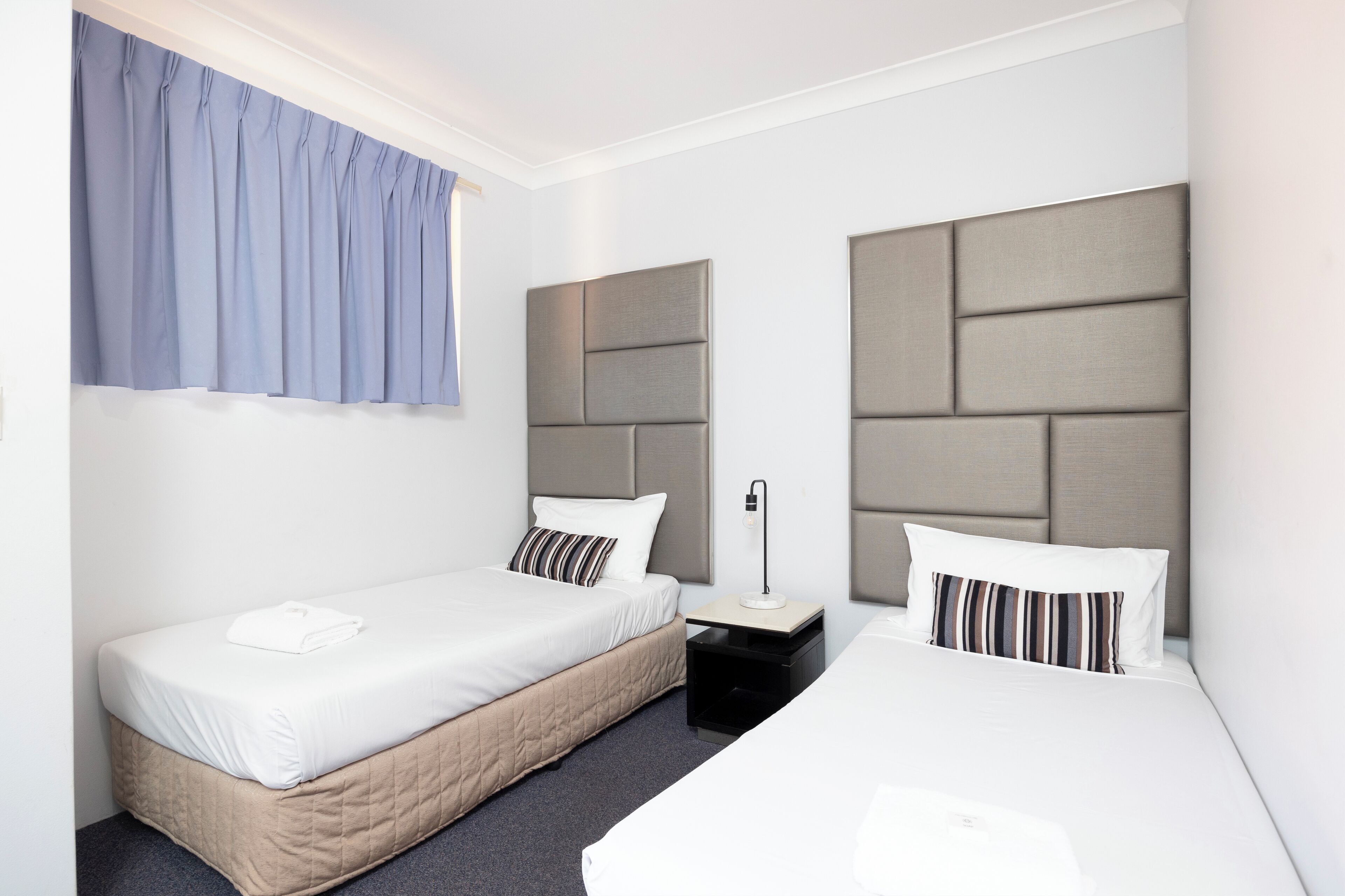 A hotel room with two beds, a nightstand, a lamp, and a window with curtains.