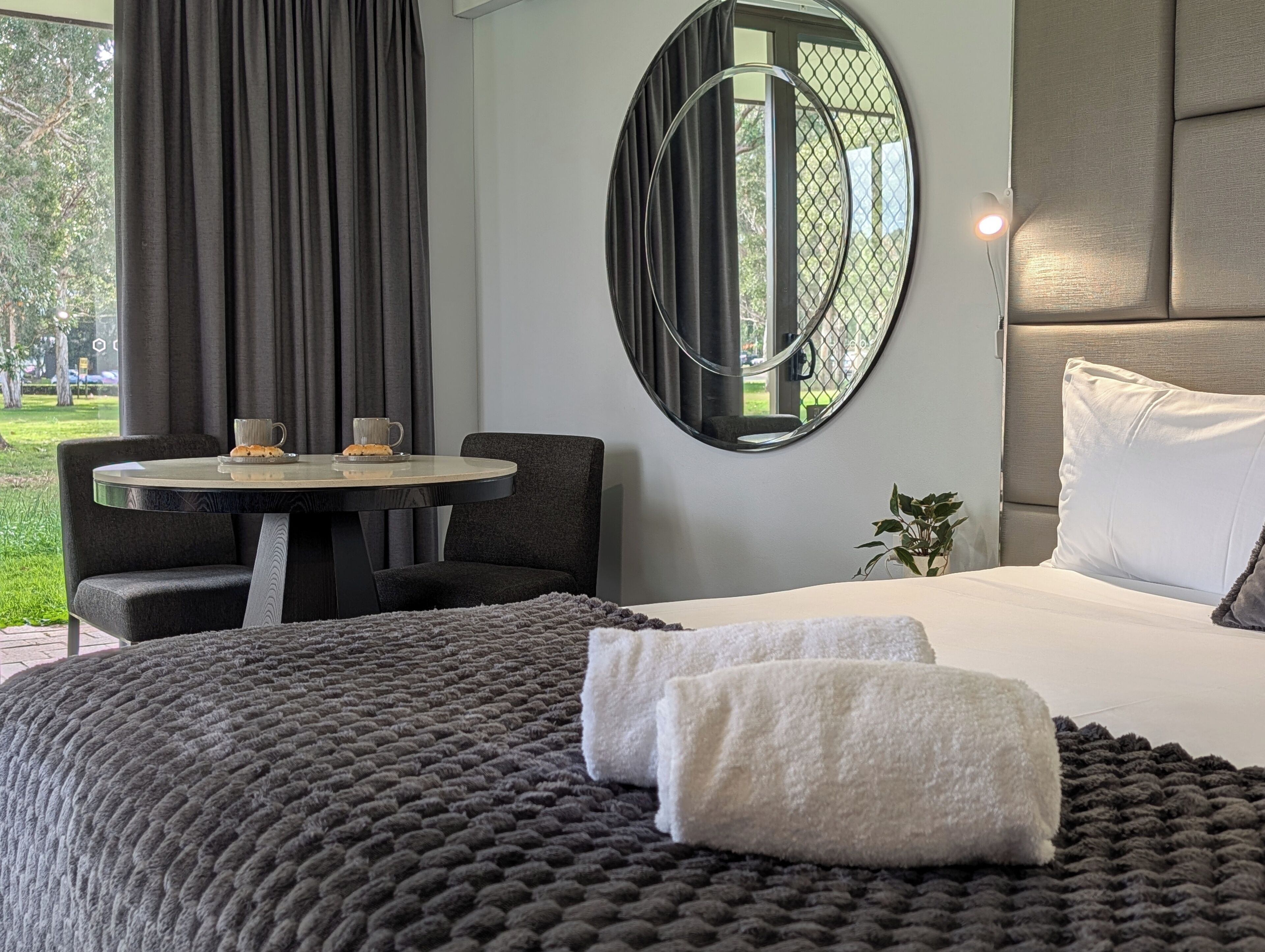 Een hotelkamer met een bed, een ronde spiegel, een tafeltje met twee kopjes, een stoel en uitzicht op de omgeving.