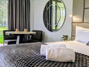 Habitación de hotel con una cama, un espejo redondo, una mesa y dos sillas, y vistas a zonas verdes al exterior.