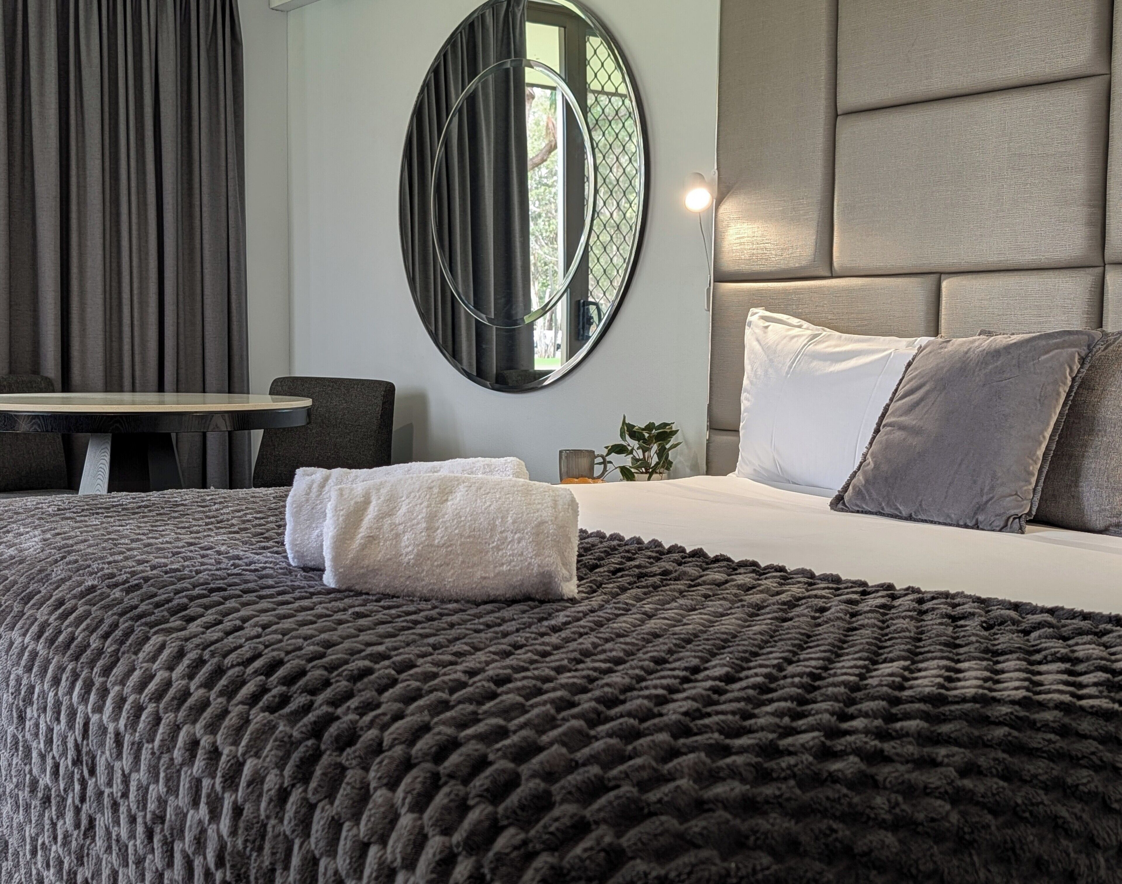 A modern hotel room with a large bed, a round mirror, a desk, and a chair.