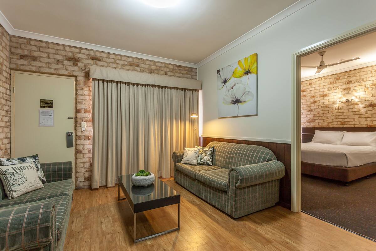 A room with a brick wall, a green plaid sofa, a wooden coffee table, a bed with a wooden headboard, and a painting of flowers on the wall.