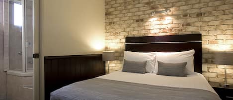 Una habitación de hotel con pared de ladrillo, una cama grande con cabecera, dos lámparas de noche y una puerta que da a una zona de ducha.