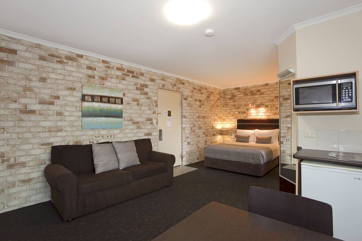 A compact hotel room with a brick-patterned wall, a sofa, a bed, a small kitchenette, and a painting.