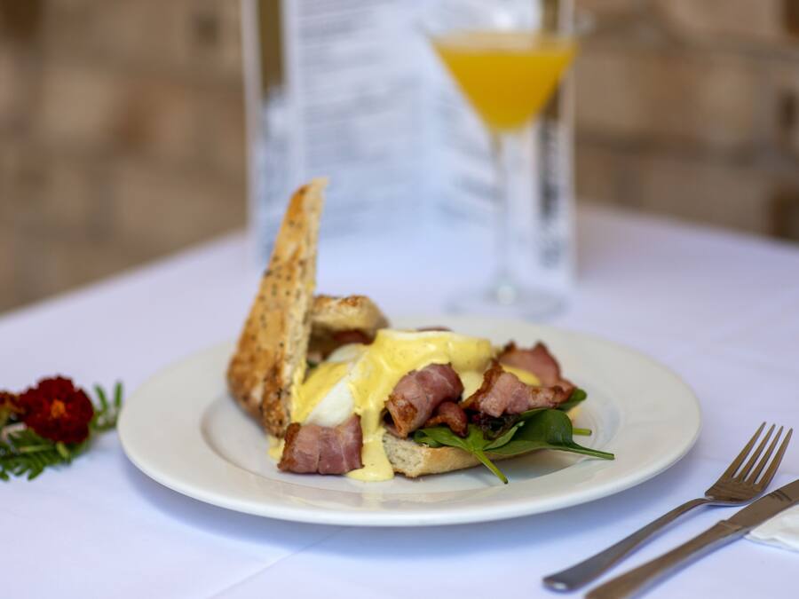 Une assiette de petit-déjeuner avec des œufs, du bacon et des légumes verts, le tout servi sur une assiette blanche, un verre de jus d’orange et une fleur sur la table.
