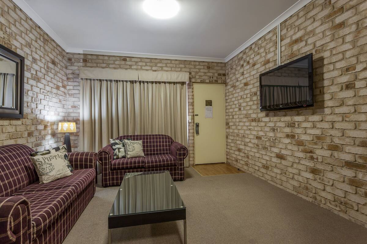 A living room with a brick wall, two plaid sofas, a coffee table, a TV mounted on the wall, and a door with a light switch.