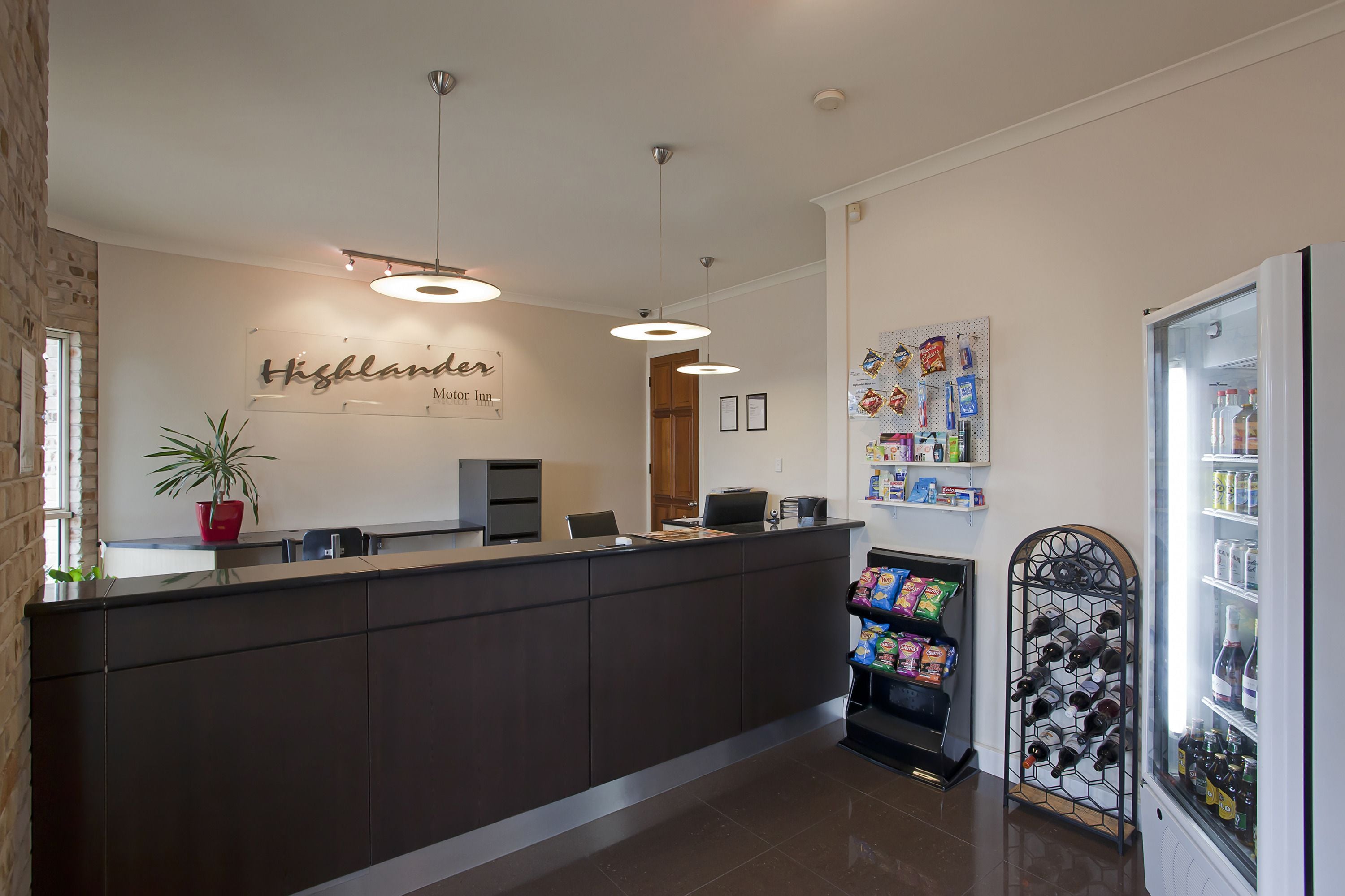 A reception desk with a sign reading "Highlander Motor Inn," a potted plant, a wine cooler, and a wall with various items.