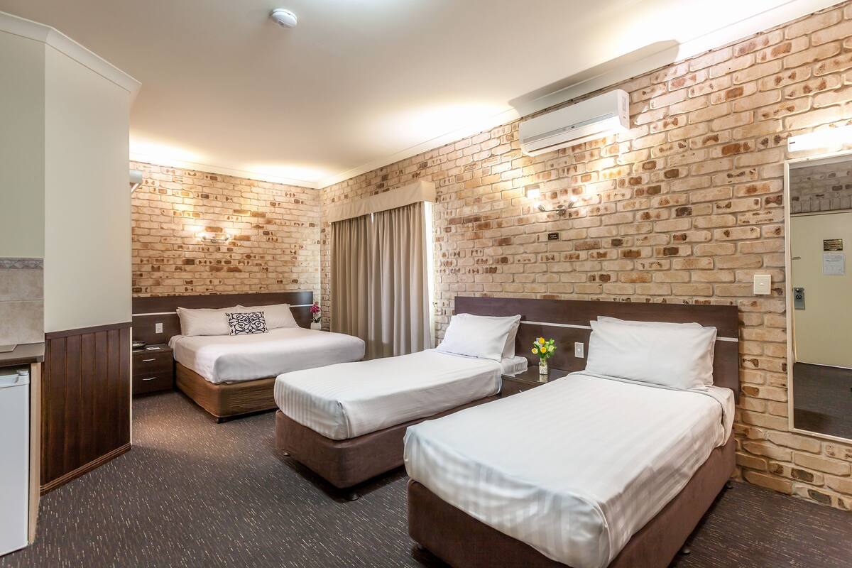 A hotel room with three beds, a wooden headboard, a brick wall, and an air conditioning unit.