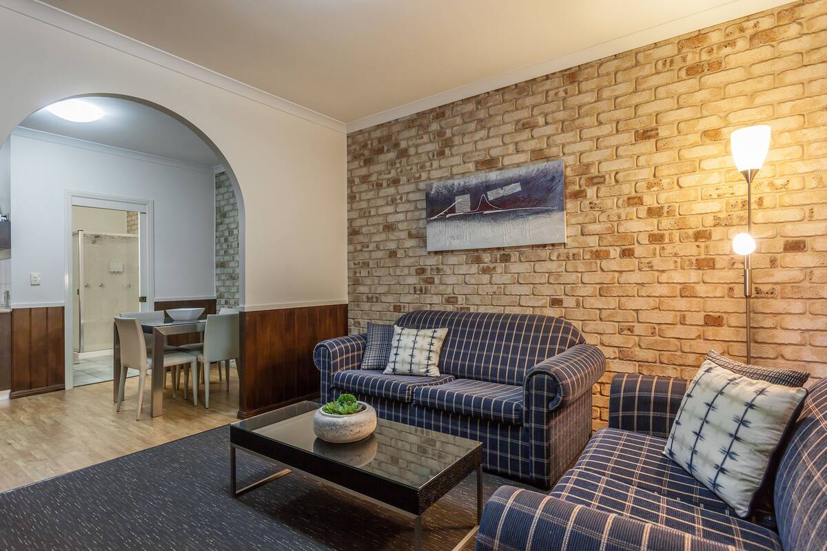 A living room with a brick wall, a sofa, a coffee table, and a dining area with chairs and a table.