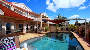 A swimming pool with a covered patio area, a table with a newspaper and a cup, and a building with a red roof and brick walls.
