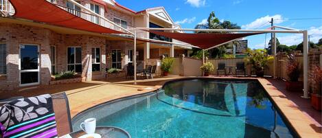 A swimming pool with a covered patio area, a table with a newspaper and a cup, and a building with a red roof and brick walls.