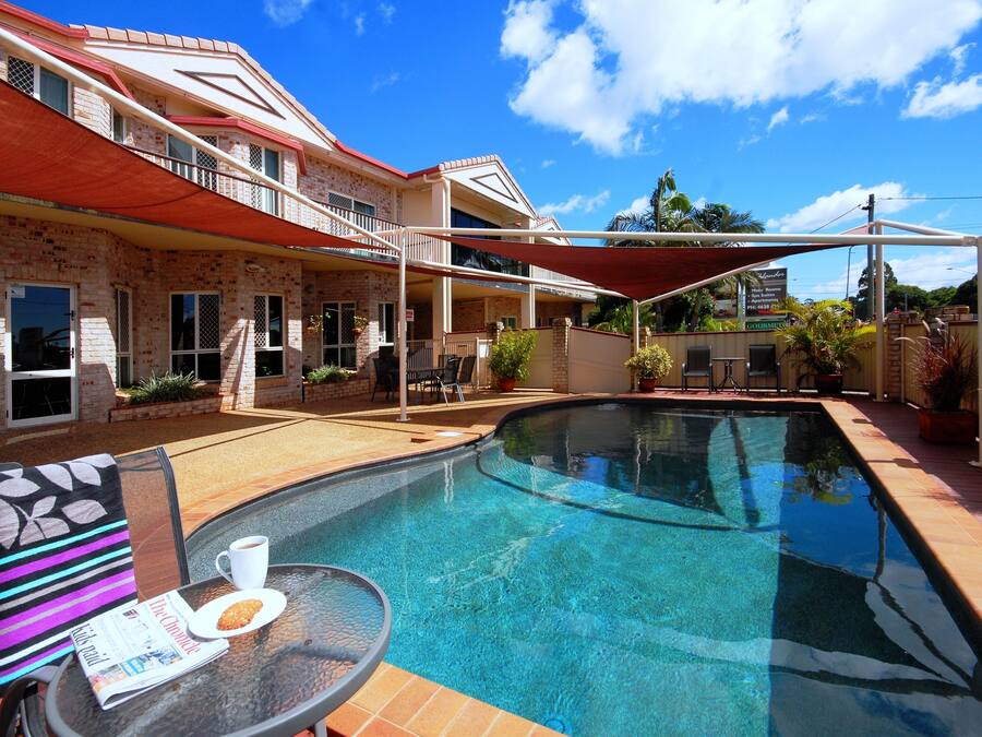 A swimming pool with a covered patio area, a table with a newspaper and a cup, and a building with a red roof and brick walls.