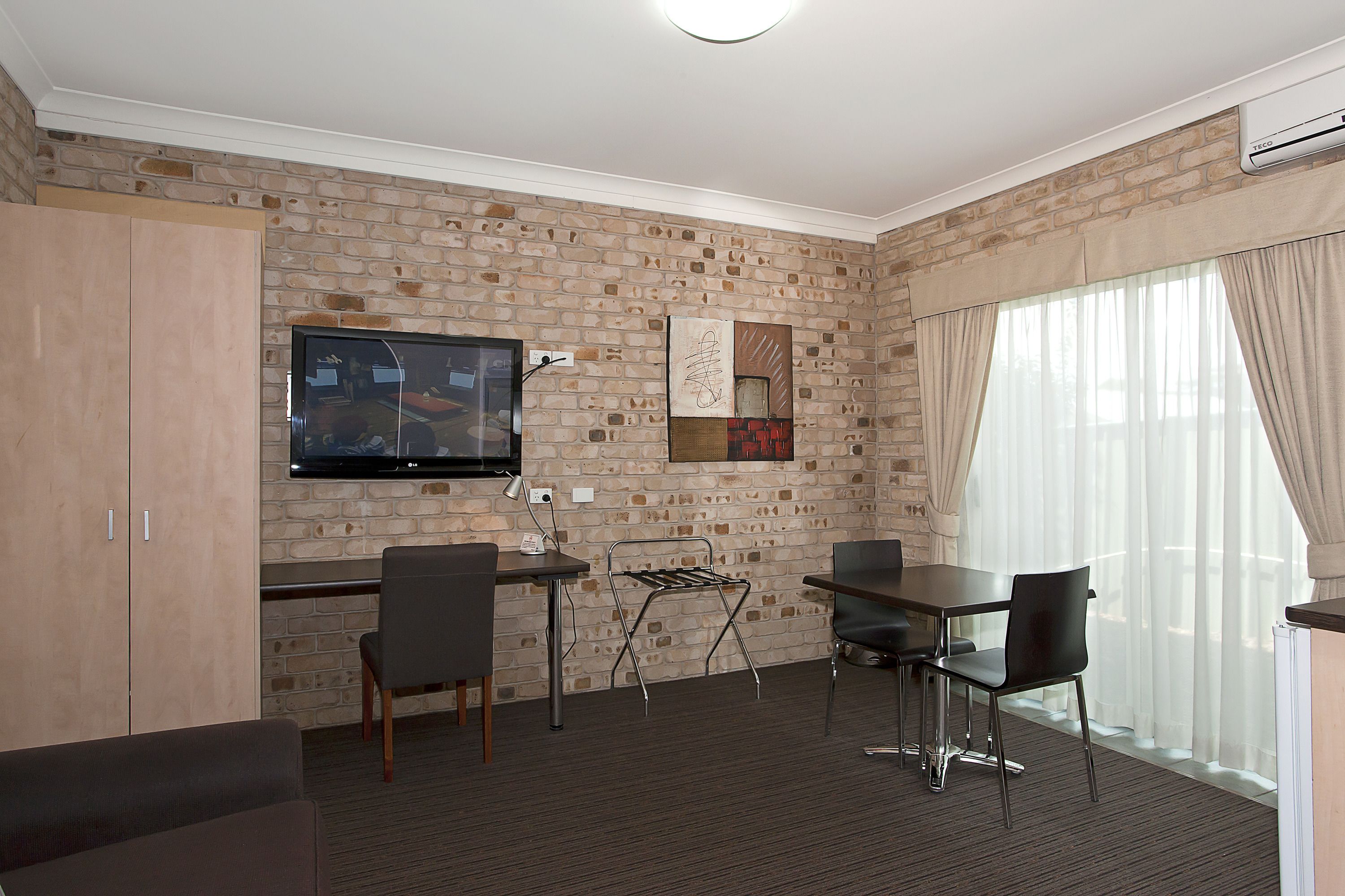 Een moderne hotelkamer met een bakstenen muur, een flatscreen televisie, een zithoek met een tafel en stoelen, en een groot raam met gordijnen.