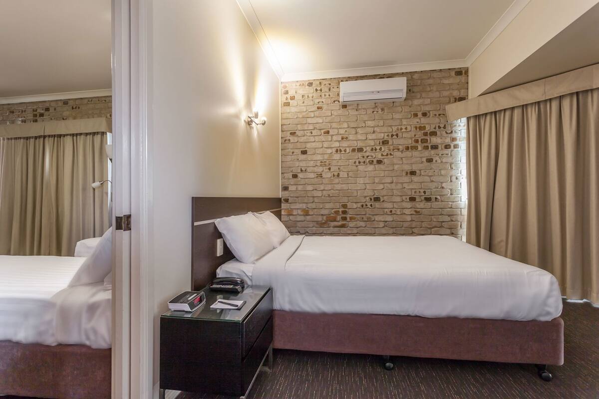 A hotel room with a brick-patterned wall, a bed, a nightstand with a phone and remote, and a window with curtains.
